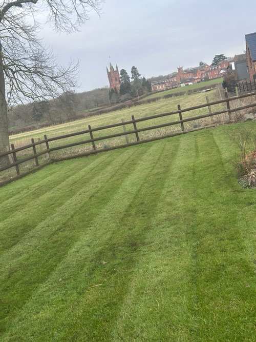 a well-maintained green lawn with striped patterns featuring a wooden fence and a distant view of a building with a tower surrounded by trees showcasing natural beauty in the landscape with five lines of freshly cut grass